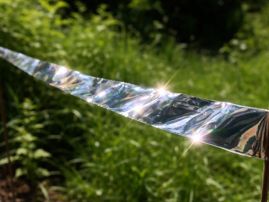Close-up of reflective mylar tape fluttering in sunlight, flashing bright highlights against greenery.
